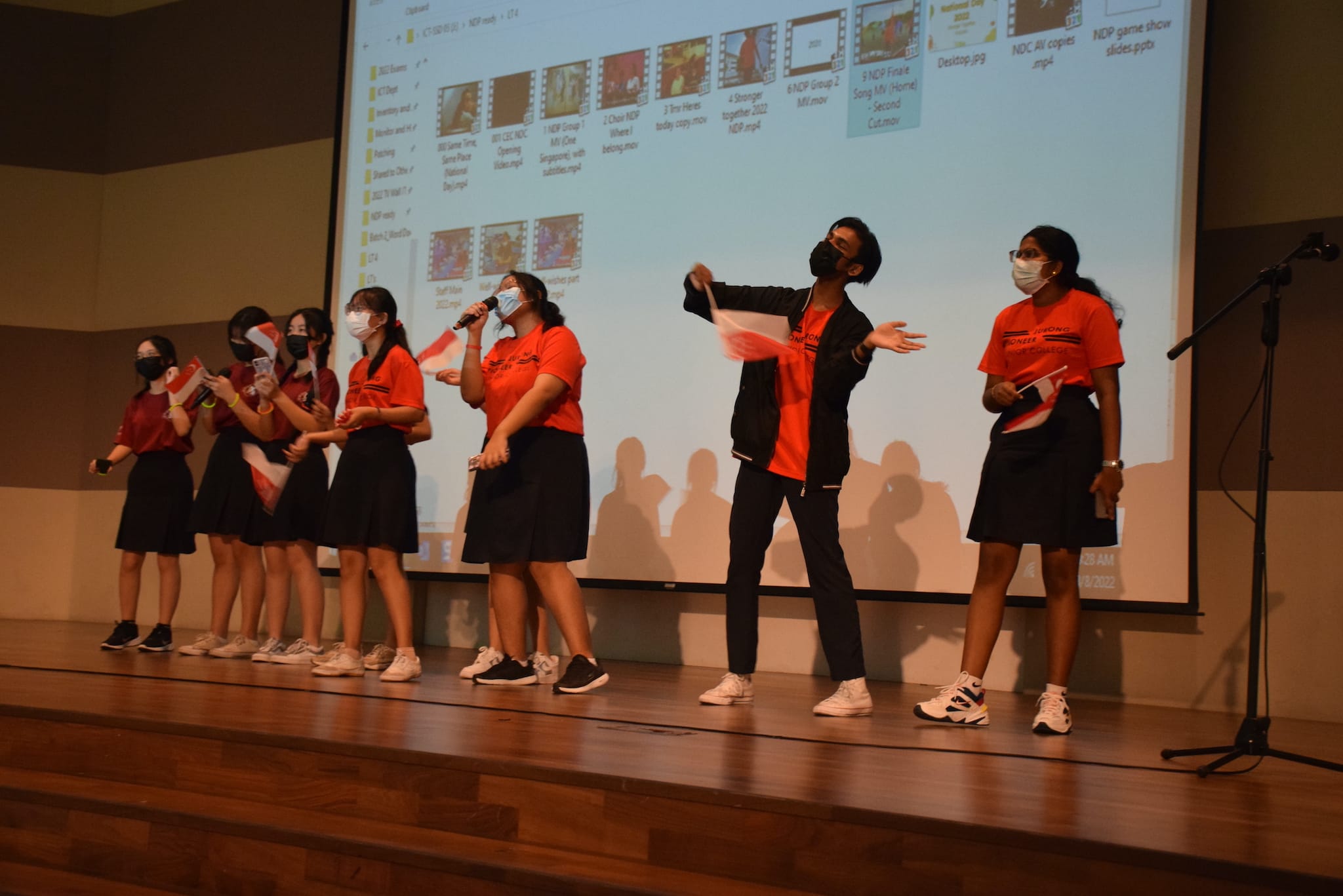 Students in red shirts and face masks perform on a wooden stage in front of a projected computer screen.